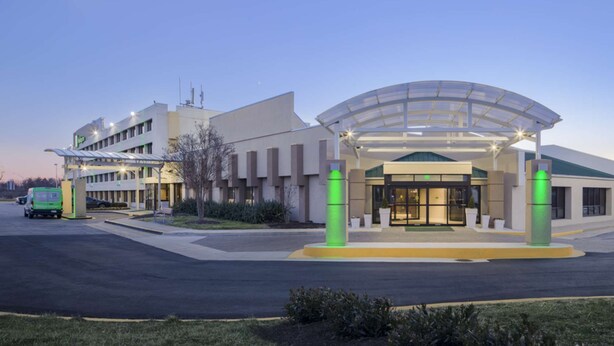 A modern hotel entrance with a covered driveway, a parking area, and a green vehicle. - Stay Near UMD | Wi‑Fi, Pool, and Restaurant (College Park)