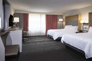 Habitación de hotel con dos camas, un escritorio con silla, un televisor y un amplio ventanal con cortinas.