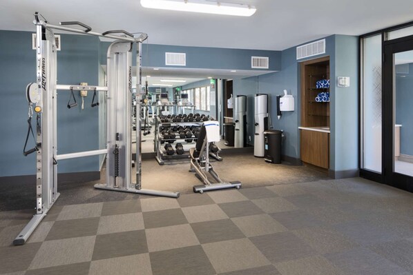 Un gimnasio con varios equipos de ejercicio, un soporte para mancuernas en la pared y un basurero.