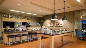 Un área de desayuno de hotel con barra de buffet, taburetes de bar y una mesa de comedor.