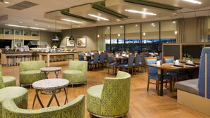 En rummelig spisesal på hotel med flere siddepladser, herunder runde borde og stole, et barområde og udsigt over bybilledet gennem store vinduer.