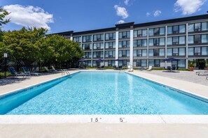 Una piscina rectangular con superficie de agua azul cristalina, rodeada de sillas de descanso y sombrillas, con un edificio moderno al fondo.