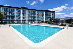 Una piscina rectangular con borde de azulejos, rodeada de sillas de descanso y sombrillas, con un edificio de varios pisos al fondo.