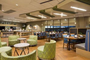 Un amplio comedor de hotel con diversas opciones de asientos, incluyendo mesas redondas y sillas, una zona de bar y vistas a la ciudad a través de grandes ventanales.