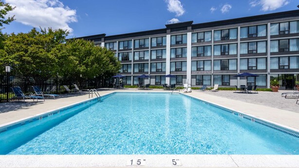 En rektangulær swimmingpool med krystalklart, blåt vand, omgivet af liggestole og parasoller, med en moderne bygning i baggrunden.
