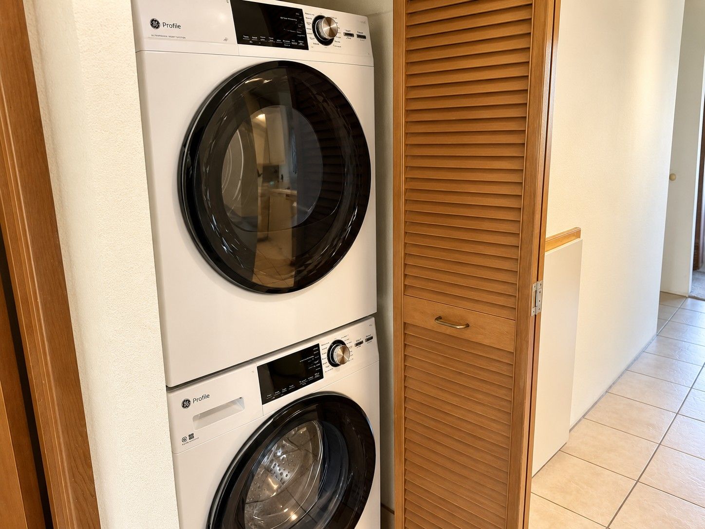 A front-loading washer and dryer unit with a control panel, located in a hallway with a wooden door and tiled flooring.