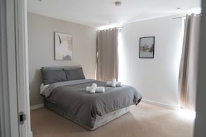 A bedroom with a bed, two pillows, a headboard, a wall-mounted artwork, a window with curtains, and a carpeted floor. - Spacious Woolwich Apartment With sea View (London)