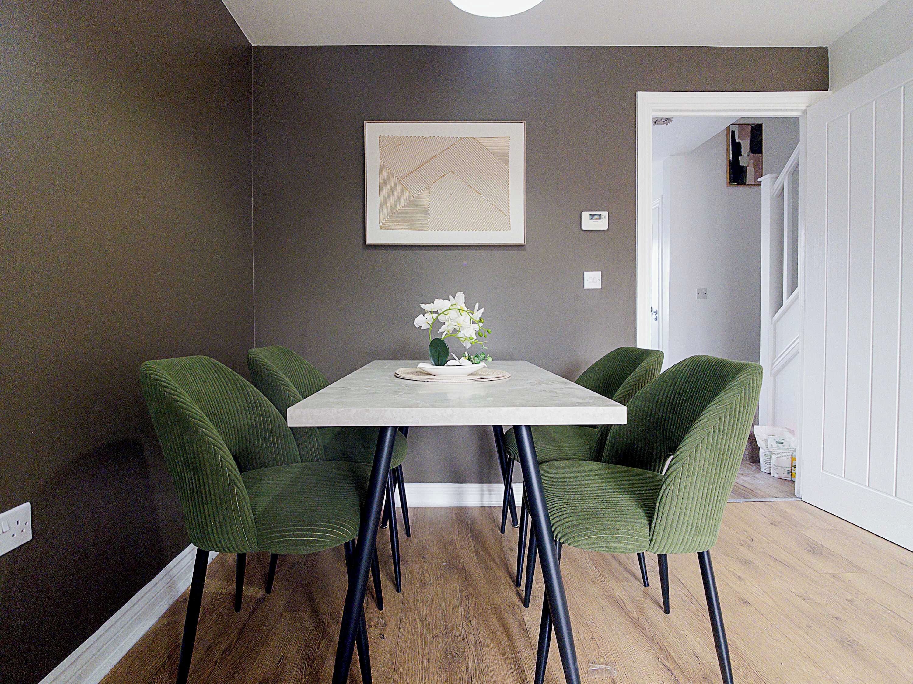 A dining area with a rectangular table, four green upholstered chairs, a white vase with flowers, and a framed artwork on the wall.