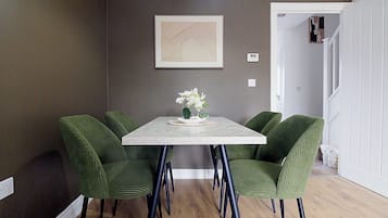 A dining area with a rectangular table, four green upholstered chairs, a white vase with flowers, and a framed artwork on the wall.
