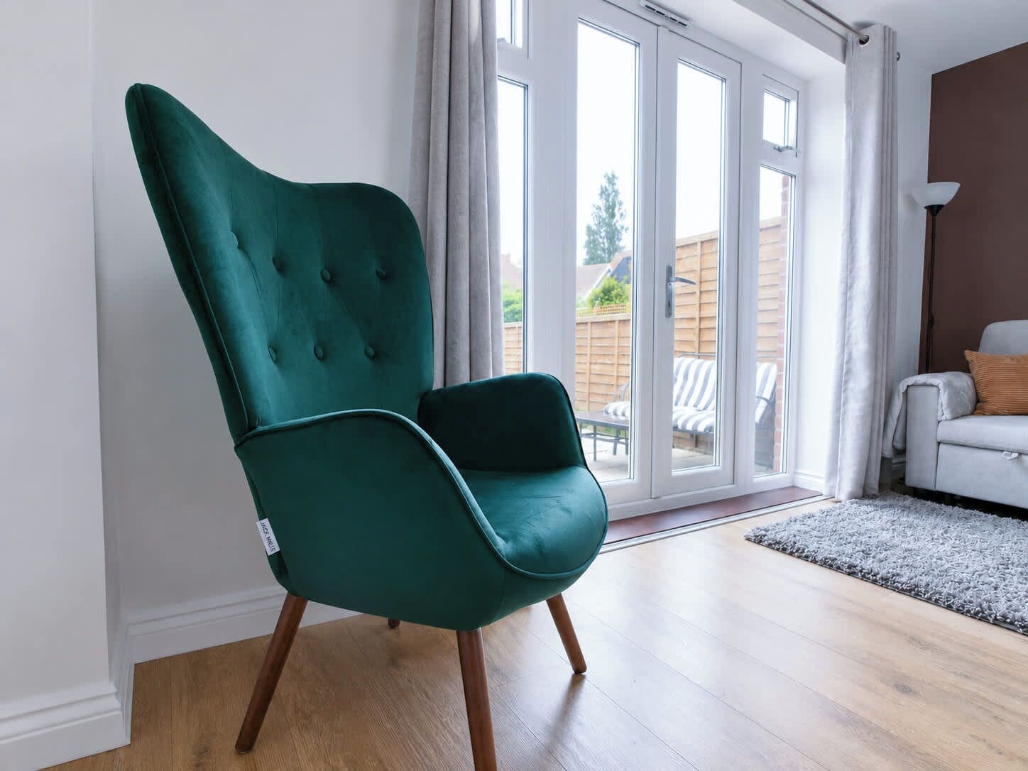 Un sillón azul verdoso con patas de madera en una habitación con suelo de madera, un cristalero corredero que da a un espacio exterior, y un sofá blanco con alfombra gris.