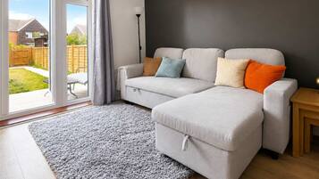 A modern living room with a grey sofa, a grey rug, a wooden side table, and a view of a backyard through a glass door.