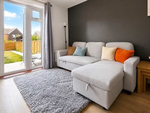 Een moderne woonkamer met een grijze bank, een grijs vloerkleed, een houten bijzettafeltje en uitzicht op een achtertuin door een glazen deur.