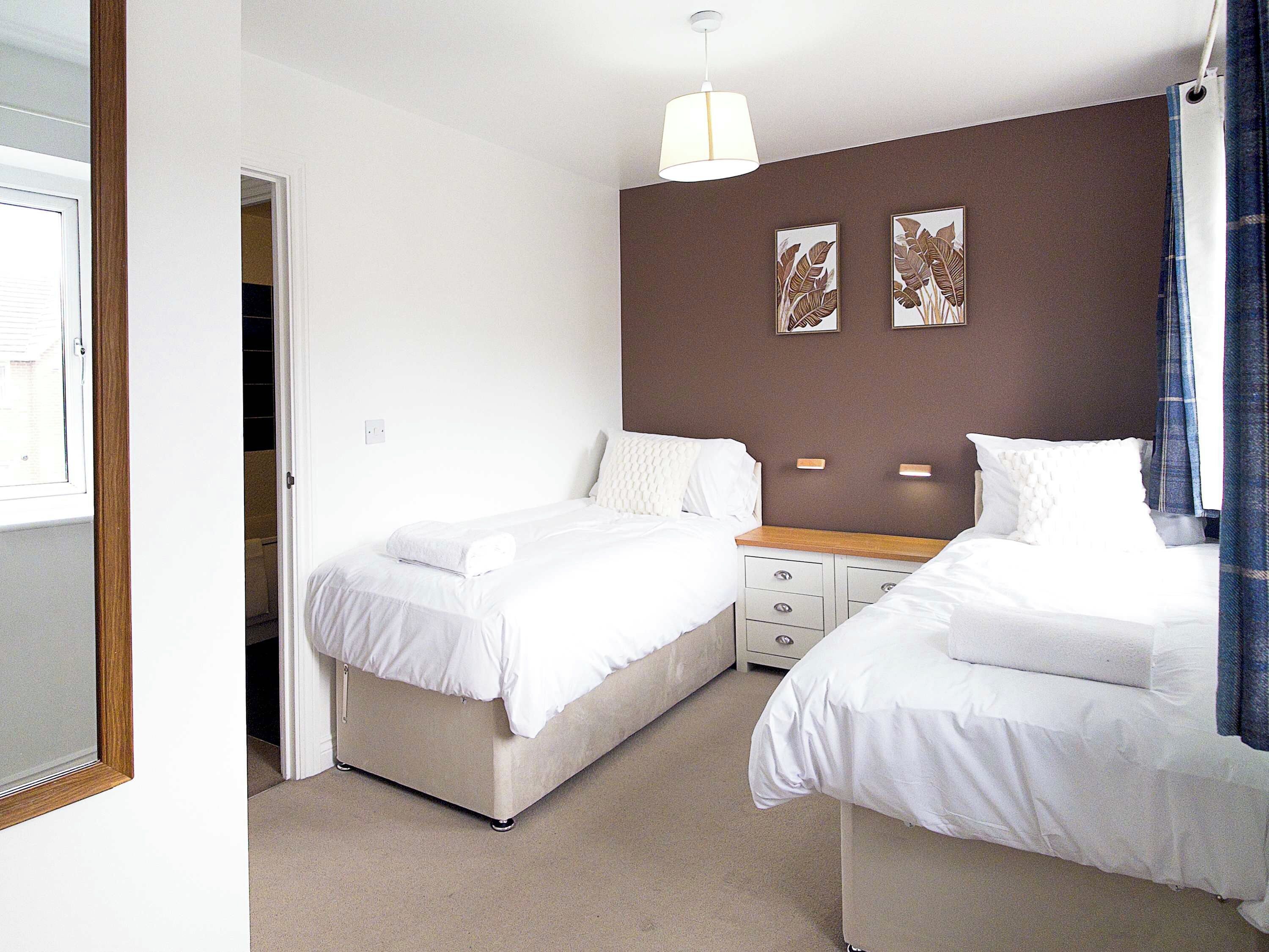 A hotel room with two beds, a wooden nightstand, a white lamp, and two wall-mounted picture frames.