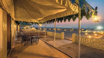 An outdoor seating area with tables and chairs, a covered patio, and a view of distant tents.