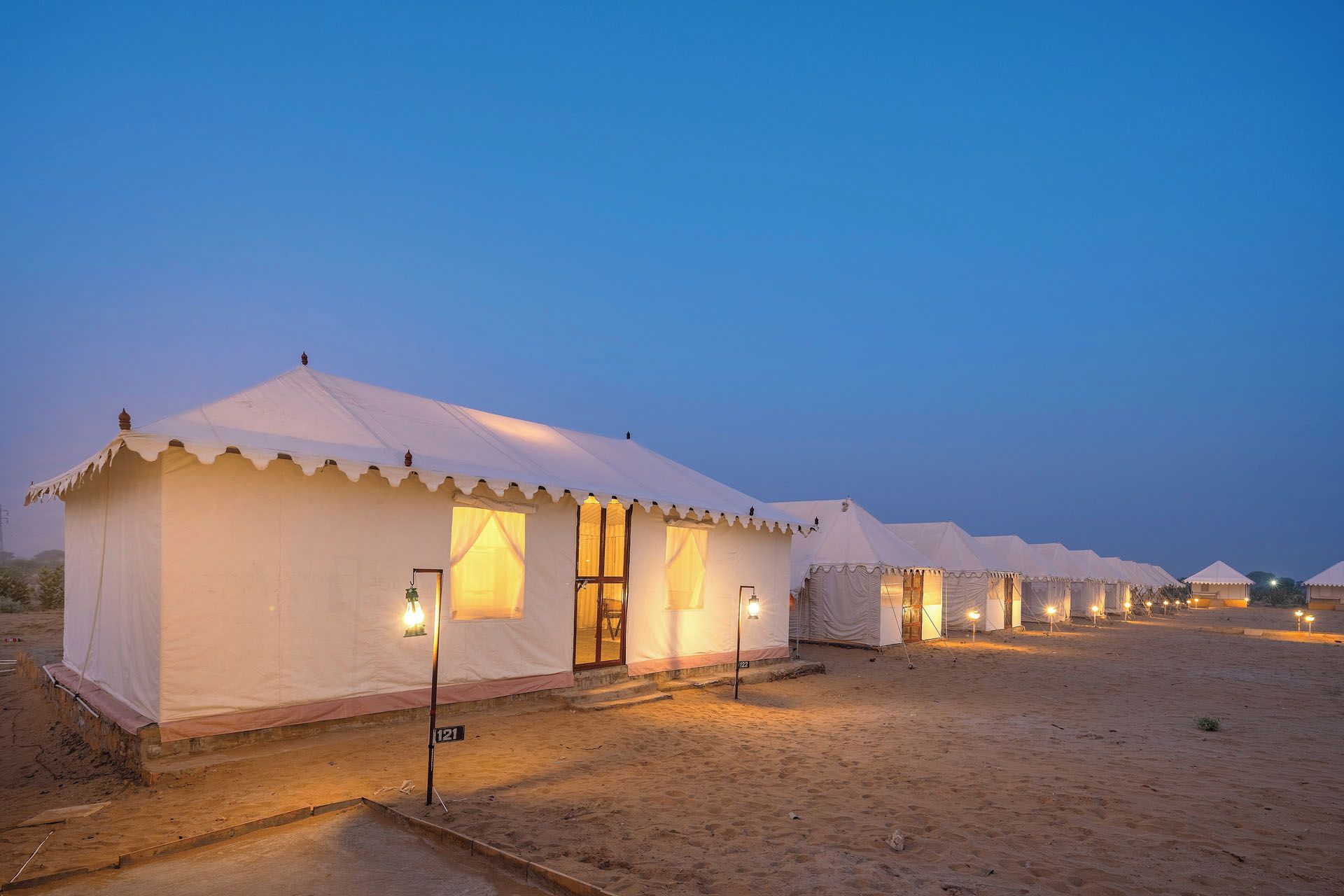A row of white tents with a flat roof, each with a small window and a light fixture outside.