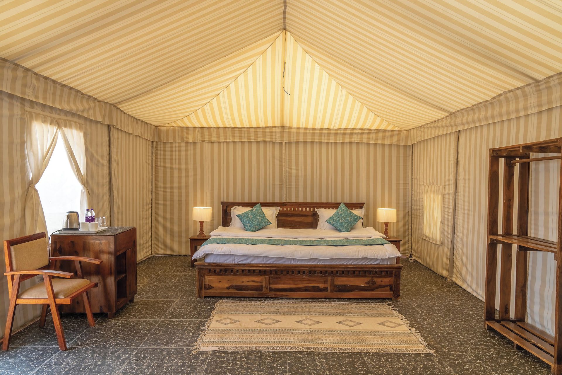 Una cama individual con cabecera de madera, dos almohadas, una mesita de noche con lámpara, una silla y un tapete estampado.