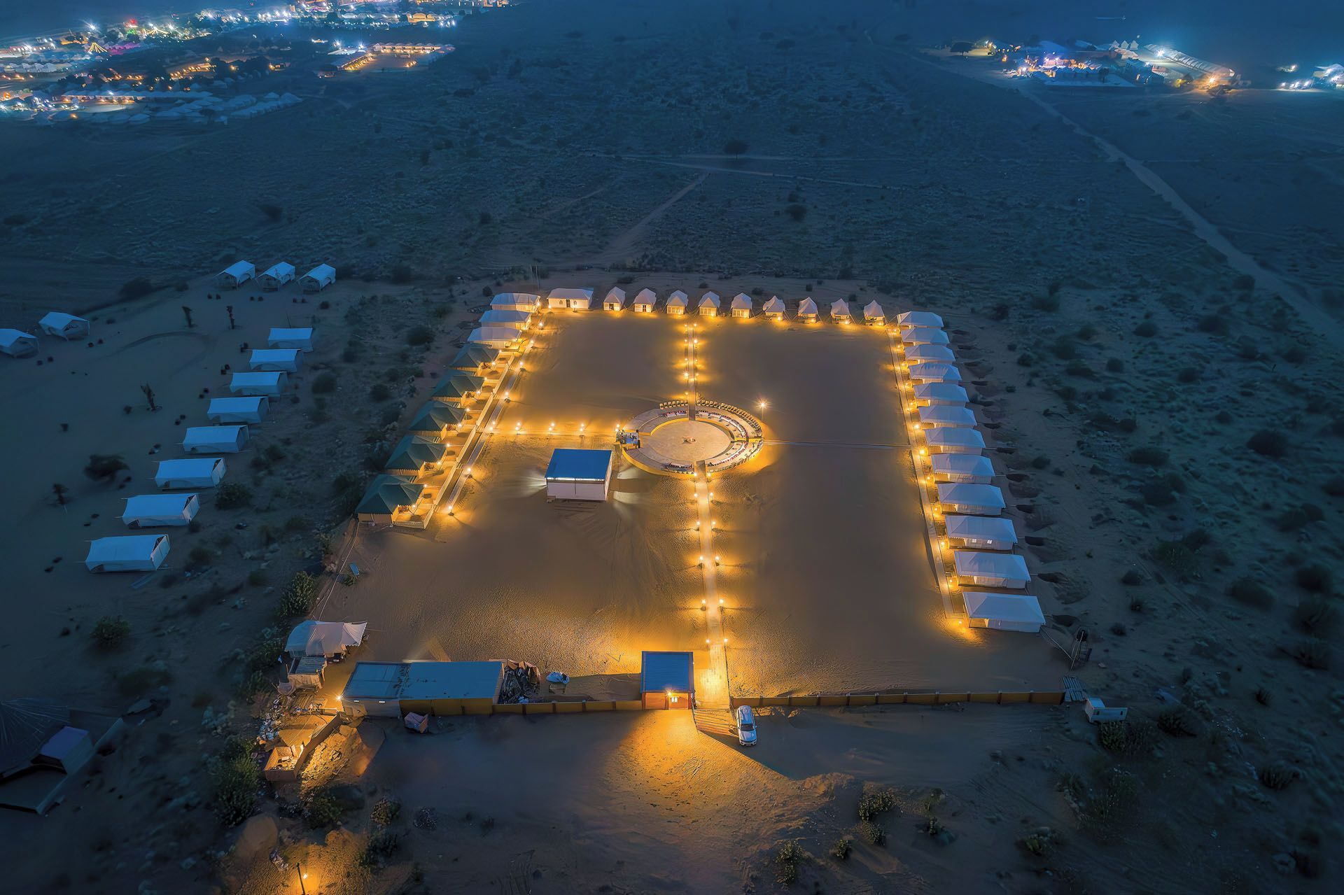 Un resort en el desierto con tiendas de campaña dispuestas en patrón cuadrado, un área circular central con luces y un camino pavimentado que lleva a la entrada.