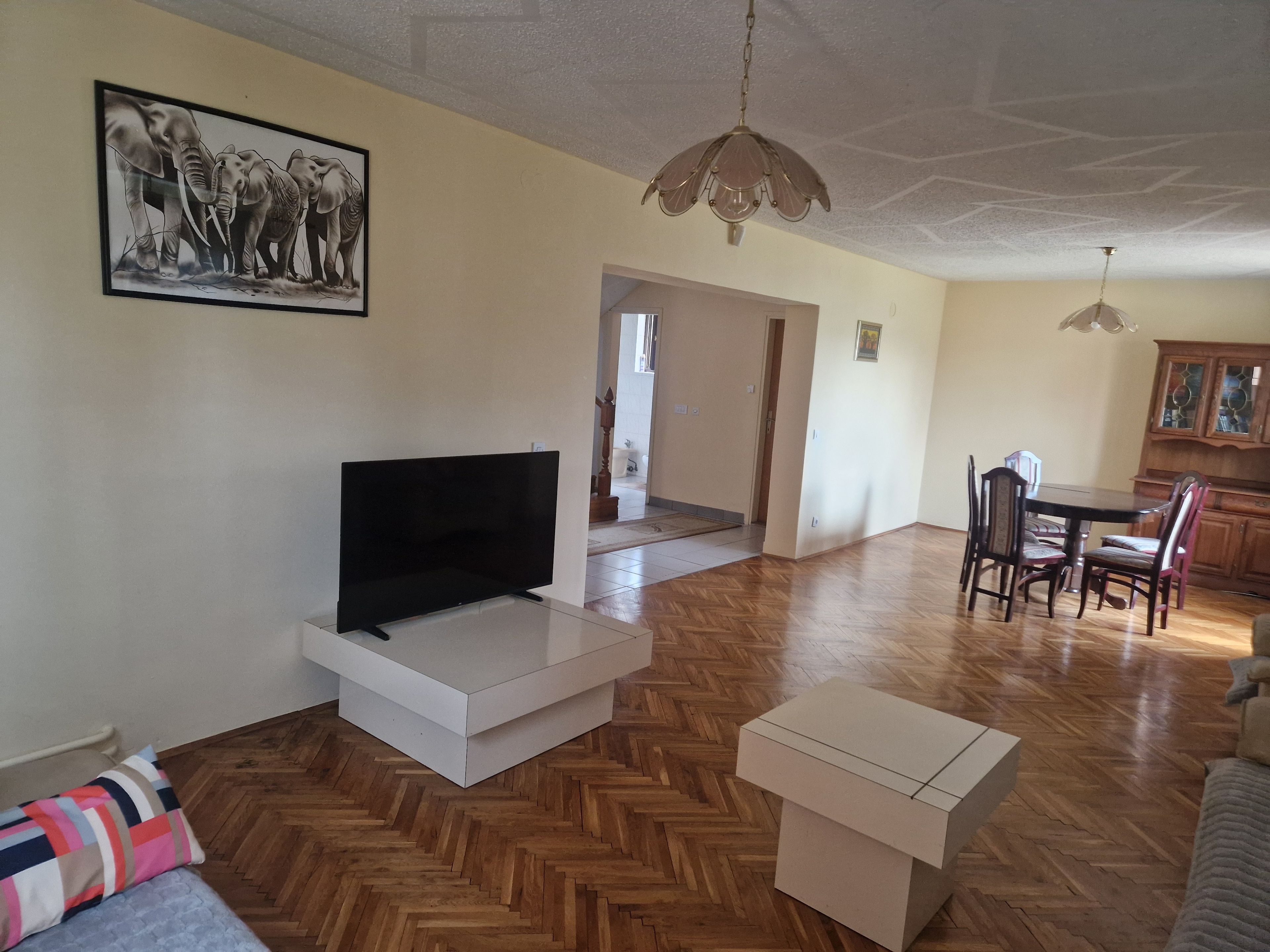 Una sala de estar con una televisión de pantalla plana, piso de madera con patrón de espiga, una zona de comedor con mesa y sillas, y un pasillo que conduce a otra habitación.