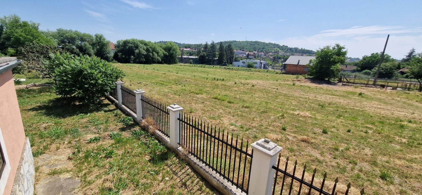 Ein eingezäunter Außenbereich mit Gras und Bäumen, ein Gebäude mit heller Fassade und ein Hügel mit Häusern im Hintergrund.