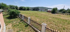 A fenced outdoor area with grass and trees, a building with a white facade, and a hill with houses in the background.