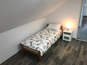 Una habitación pequeña con una cama individual, cabecera de madera, colcha estampada, mesita de noche con lámpara y vista a un pasillo.
