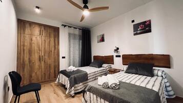 A room with two beds, a wooden wardrobe, a ceiling fan, and a small table.
