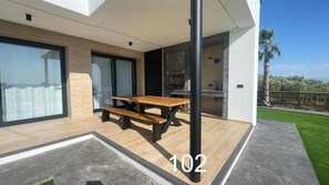 A modern outdoor dining area with a wooden table and benches, a kitchen area, and a view of a green lawn and palm trees.