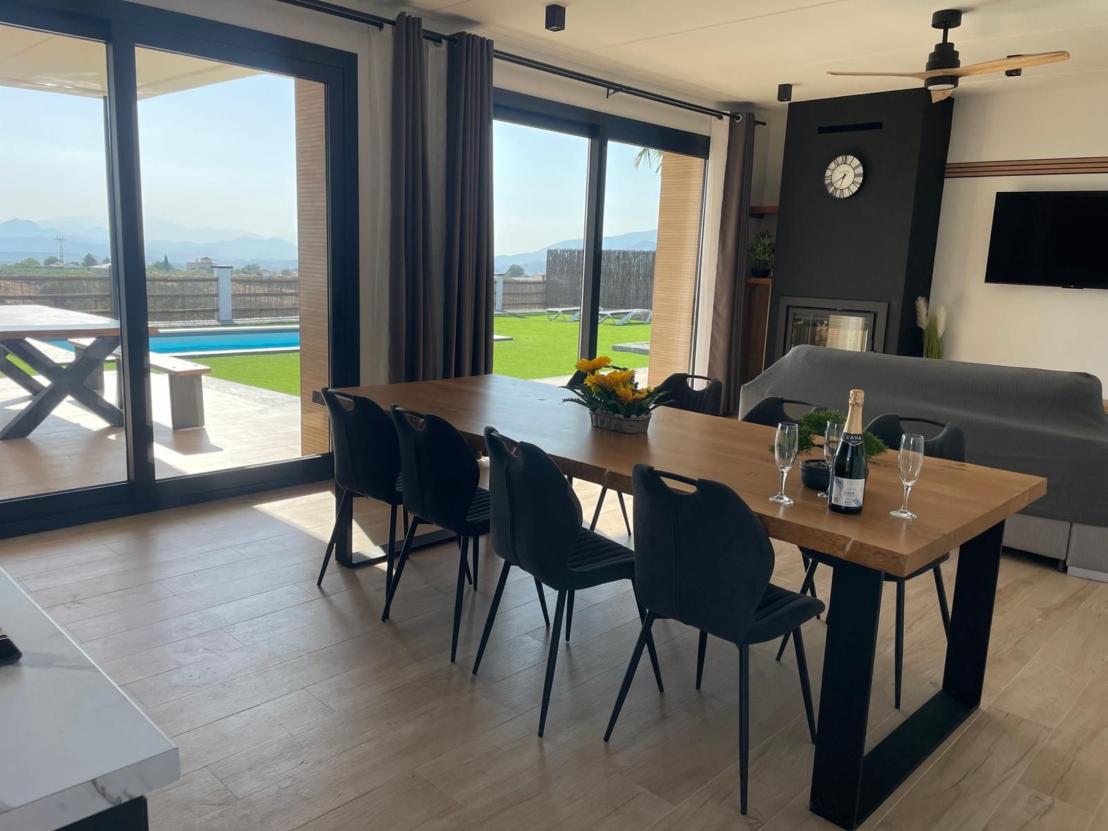 Un comedor moderno con una mesa de madera, sillas negras y vistas a una piscina y montañas.