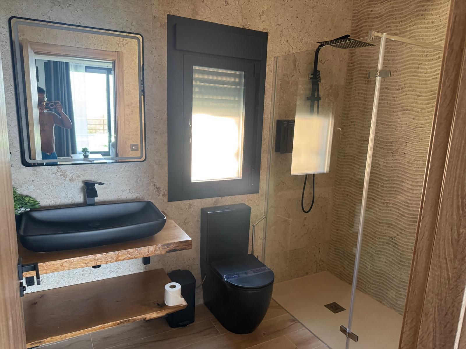 Un baño con tocador de madera, un lavabo negro, inodoro y zona de ducha.