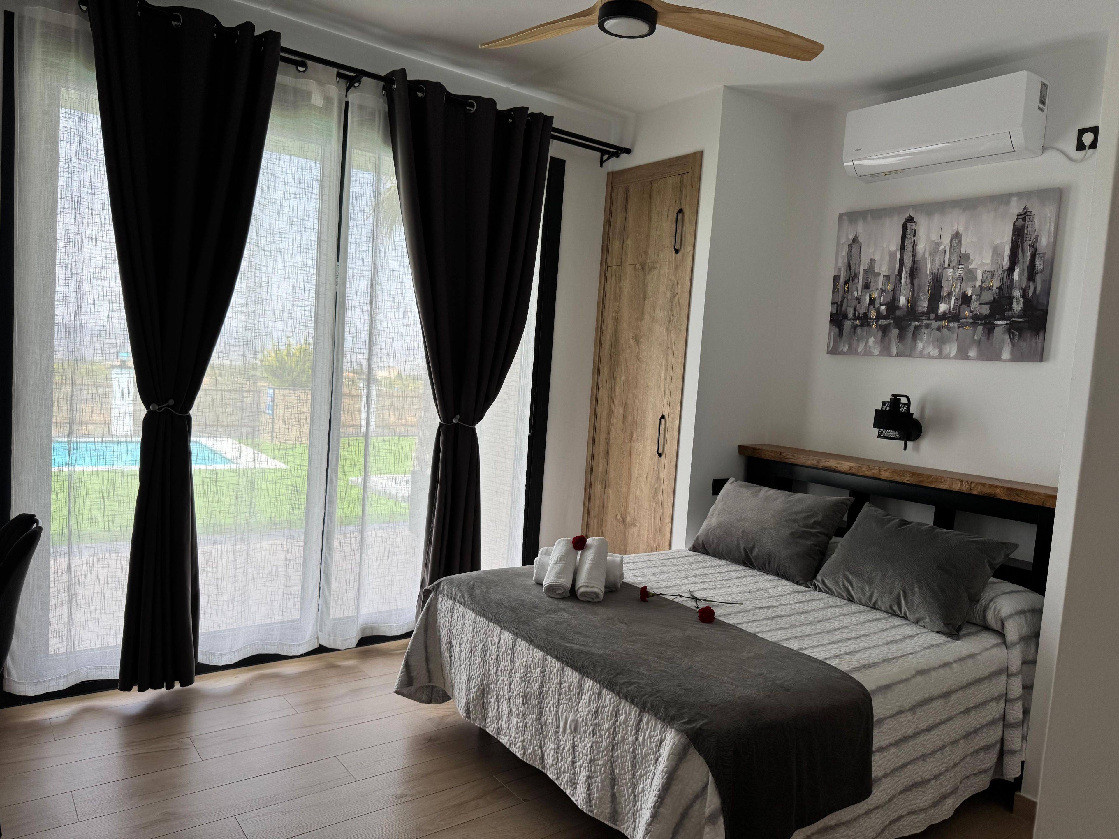 Un dormitorio con cama, cortinas, ventilador de techo, aire acondicionado, un cuadro y vista a una piscina.