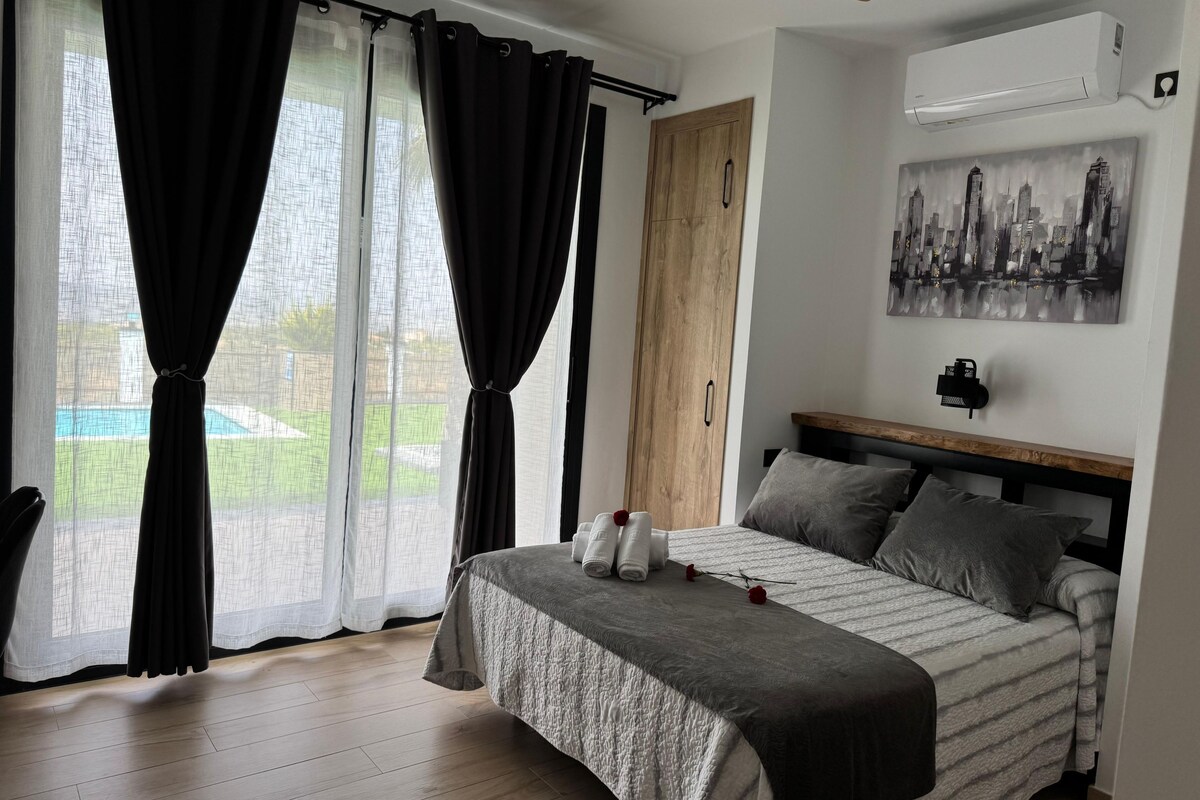 Un dormitorio con cama, cortinas, ventilador de techo, aire acondicionado, un cuadro y vista a una piscina.