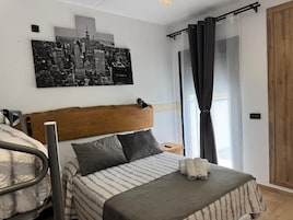 Un dormitorio con una cama, un cabecero, una pintura de paisaje urbano, una ventana con cortinas y un armario de madera.
