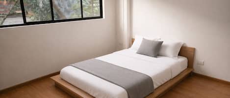 Una cama individual con estructura de madera, ropa de cama blanca y una manta gris.