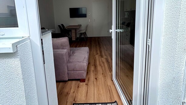 Een woonvertrek met een schuifdeur naar een balkon, een bank, een televisie en een houten vloer.