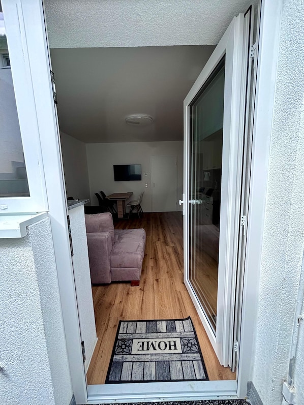 Ein Wohnraum mit einer Schiebeglastür, die auf einen Balkon führt, einer Couch, einem Fernseher und einem Holzboden.