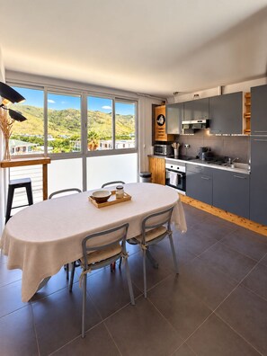 A modern kitchen with dark cabinets, a microwave, and a stove. There is a dining table with chairs and a view of a mountainous landscape through large windows. - A tropical retreat under the Saint Martin sun (Cul-de-Sac)