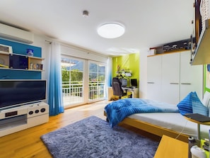A modern bedroom with a bed, a desk, a bookshelf, and a large window with a view of greenery.