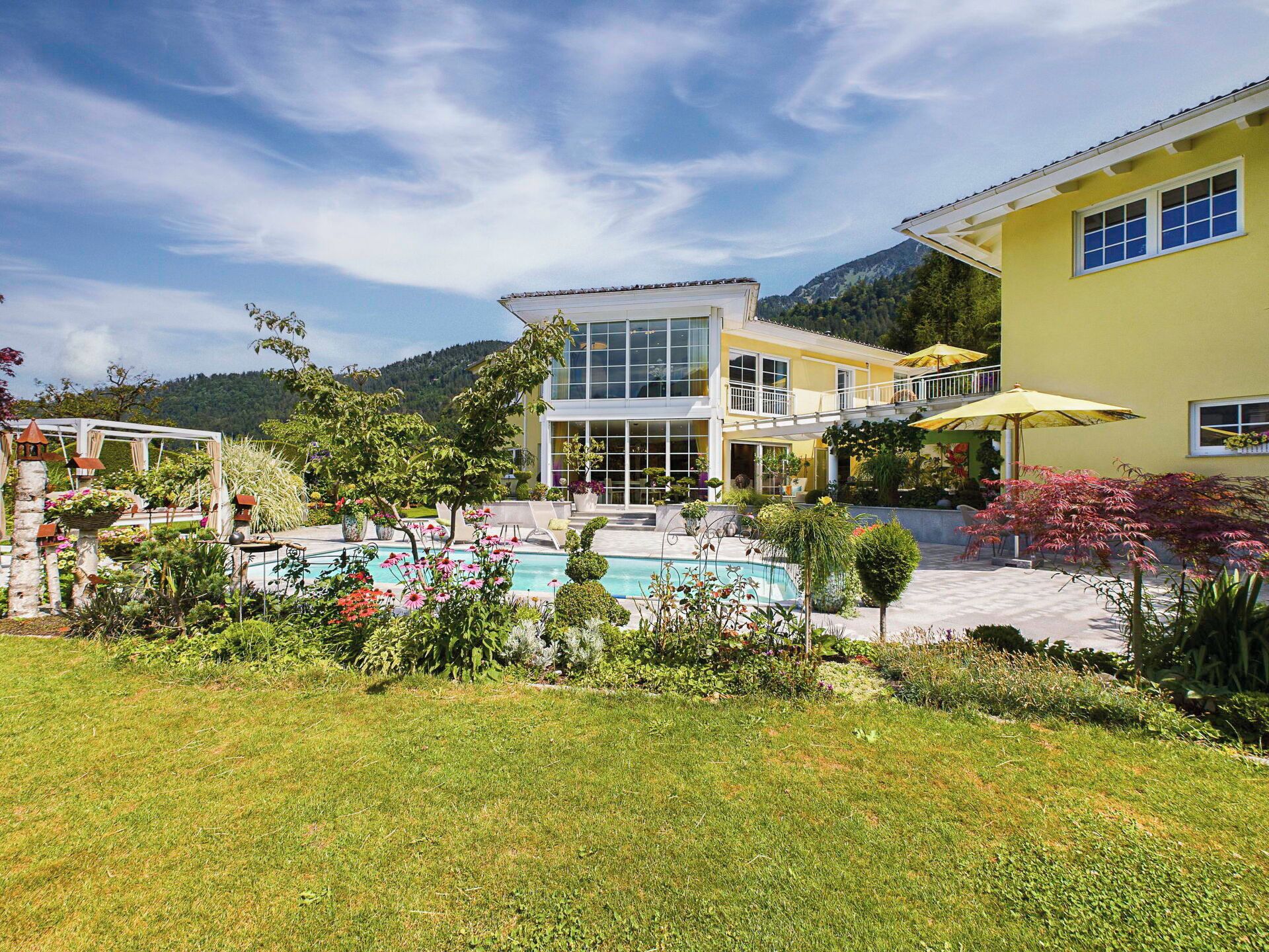 A two-story building with a swimming pool, surrounded by lush greenery and a mountainous backdrop.