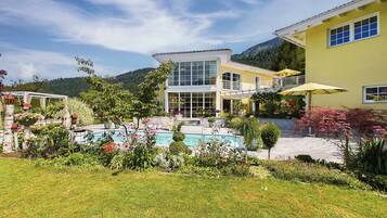 A two-story building with a swimming pool, surrounded by lush greenery and a mountainous backdrop.