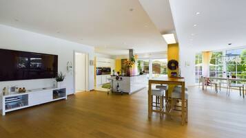 A spacious living area with a wooden floor, a central island with bar stools, a flat-screen TV, and a large window offering a view of greenery.