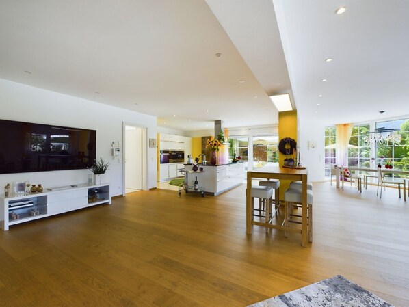A spacious living area with a wooden floor, a central island with bar stools, a flat-screen TV, and a large window offering a view of greenery.