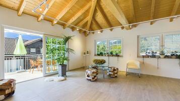 Une pièce spacieuse avec un haut plafond en bois, de grandes fenêtres et un balcon aménagé avec du mobilier d’extérieur.