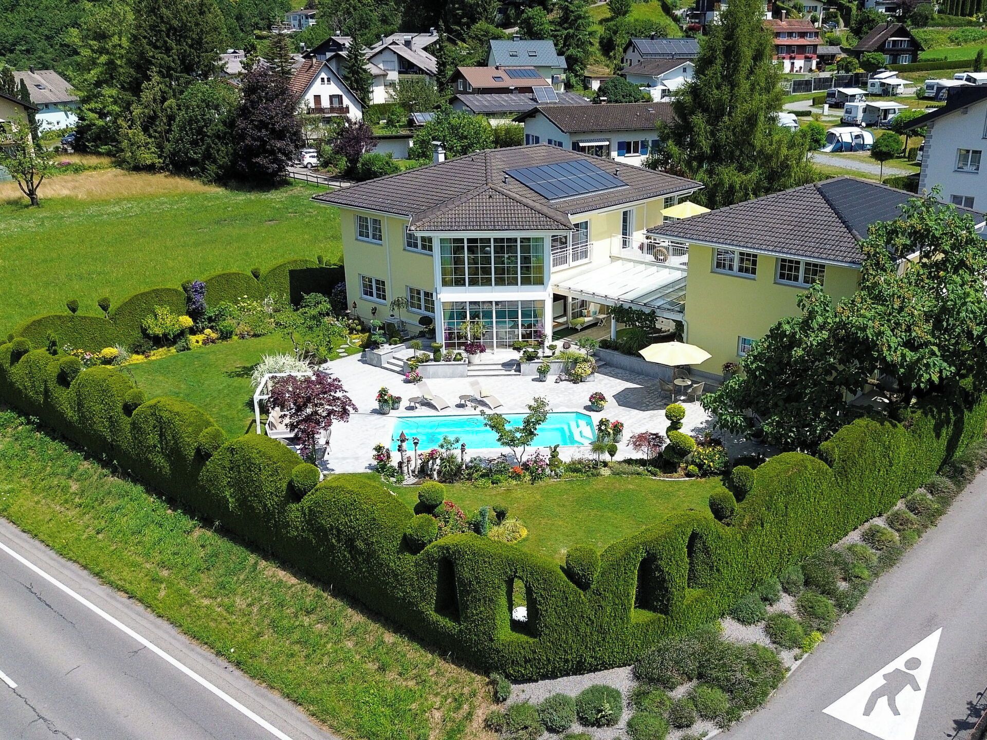 An aerial view of a residential area with a large house featuring a swimming pool, surrounded by well-maintained gardens and a road.