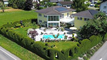 An aerial view of a residential area with a large house featuring a swimming pool, surrounded by well-maintained gardens and a road.