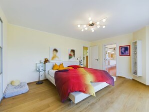 A modern bedroom with a bed, bedside tables, a nightstand, a ceiling light fixture, and a door leading to another room.