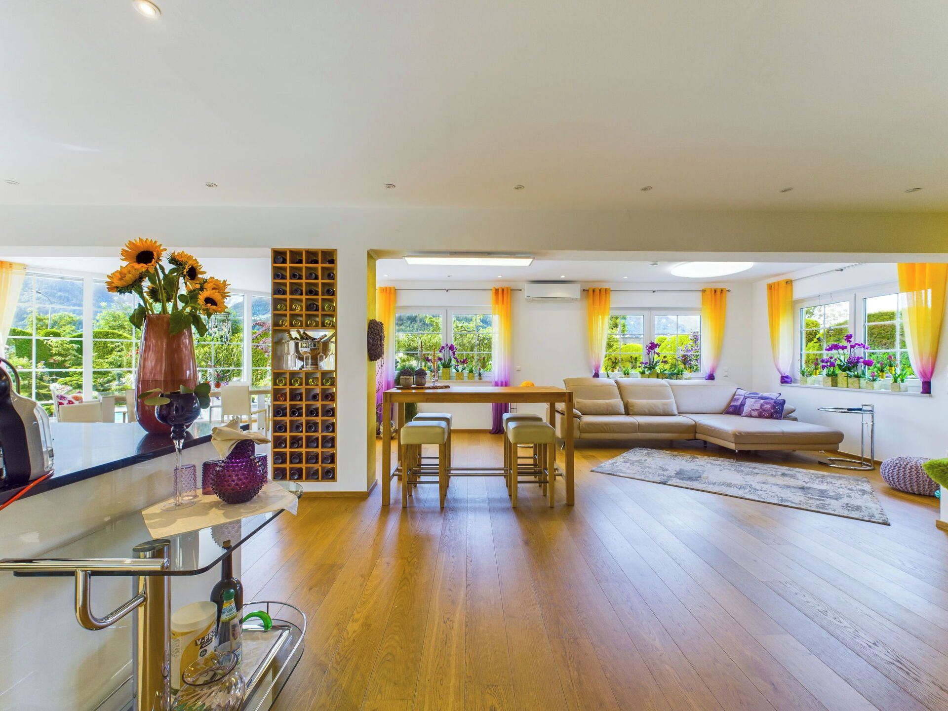 A spacious living area with a wooden floor, a sofa, a dining table with chairs, and a kitchen area with a wine rack and a vase of flowers.