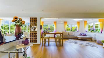 A spacious living area with a wooden floor, a sofa, a dining table with chairs, and a kitchen area with a wine rack and a vase of flowers.