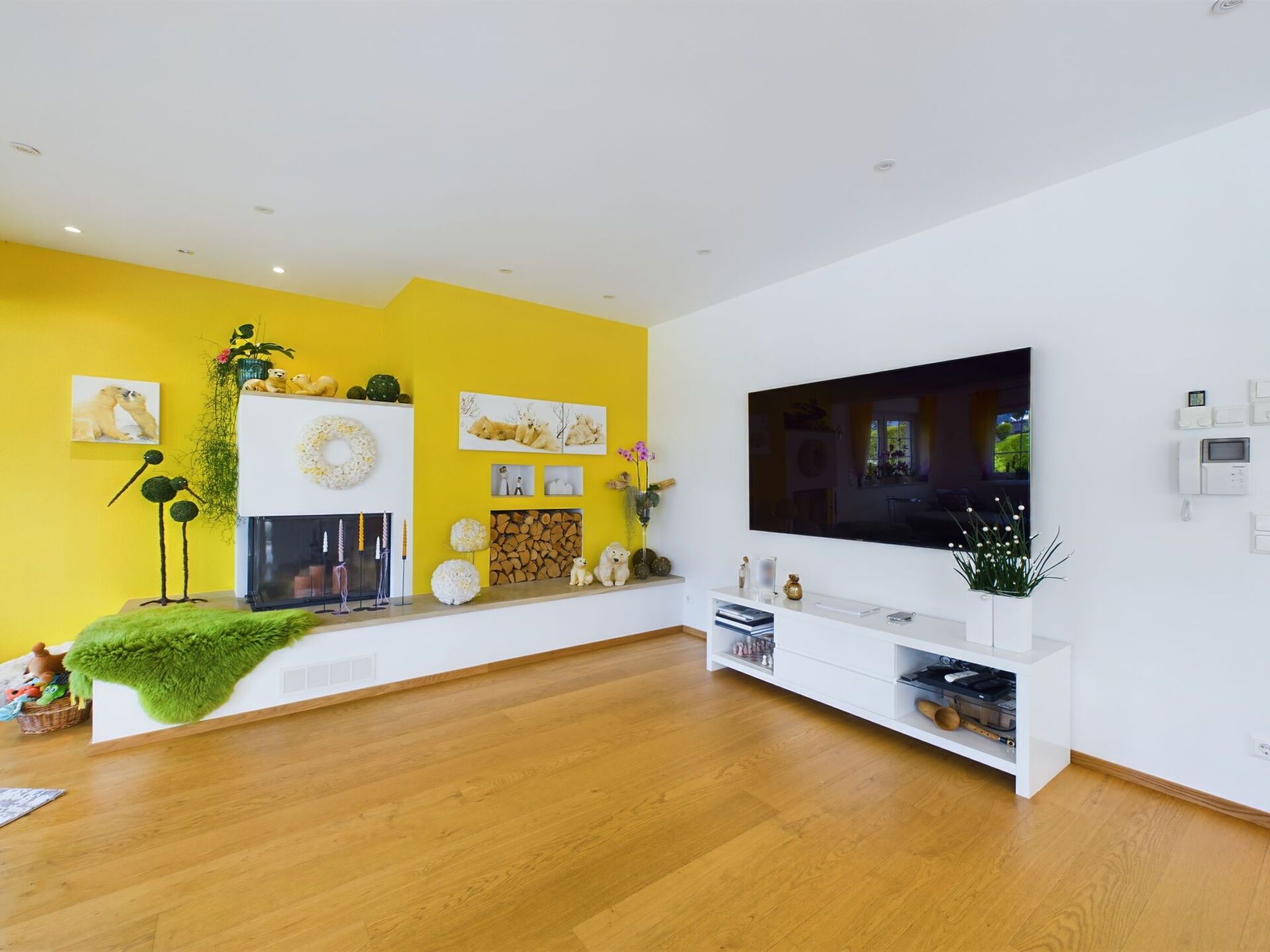 A modern living room with a large flat-screen TV, wooden flooring, a fireplace, and a bright yellow accent wall.
