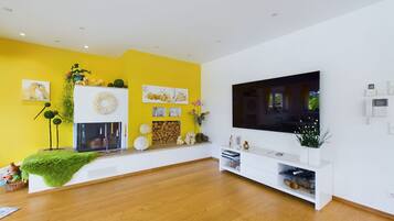 A modern living room with a large flat-screen TV, wooden flooring, a fireplace, and a bright yellow accent wall.