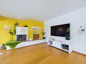 A modern living room with a large flat-screen TV, wooden flooring, a fireplace, and a bright yellow accent wall.